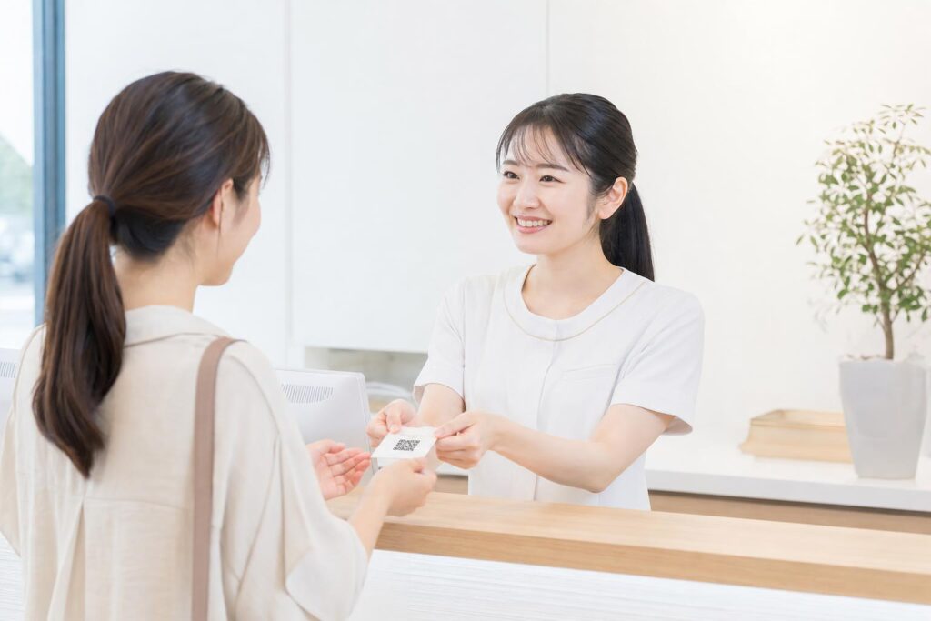クリニックの受付カウンターで、医療事務スタッフが会計時に患者へLINE友だち追加のQRコードが印刷されたカードを笑顔で手渡している場面。明るく清潔な院内、自然光